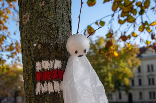 Halloween ghost decoration on tree in autumn city park with fall leaves, spooky playful outdoor seasonal atmosphere