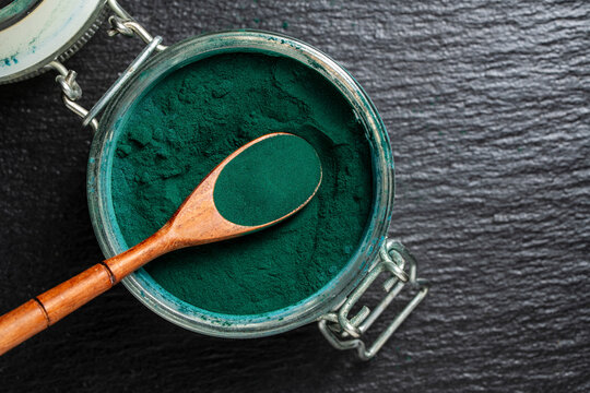 Spirulina powder in glass jar with wooden spoon, closeup. Organic green chlorella or spirulina powder inside open jar with wood spoon. Superfood supplement backdrop for healthy diet wellness and detox
