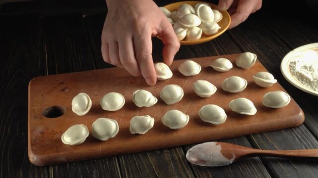 Hands arranging handmade dumplings from a bowl onto a wooden cutting board beside a spoon and flour dish, overhead view of homemade food preparation