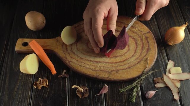Hands slicing a red beet with a kitchen knife on a wooden cutting board surrounded by vegetables, herbs, garlic, onion, carrot, potato, and mushrooms in a top view