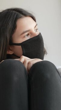 A young woman sits with her knees drawn up, wearing a black mask. The setting is indoors, highlighting the woman's focused expression and the soft lighting around her.