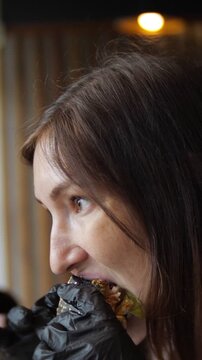 Profile view of a young woman with appetite eating a juicy burger in a cafe. She wears black gloves to eat the big sandwich and chews with pleasure, enjoying her fast food meal for lunch