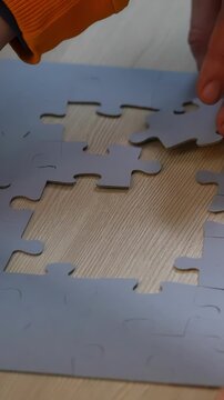 A hand is placing a puzzle piece on a table. The puzzle pieces are gray and arranged on a wooden surface, indicating an ongoing puzzle activity.