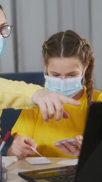 A girl wearing a mask sits at a table focused on studying. She examines study materials while a hand gestures toward her, indicating further instruction.