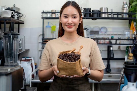 A smiling woman barista holds a burlap sack with roasted coffee beans inside her independent local caf&eacute;.