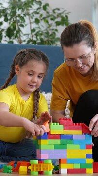 A woman and a girl are building a colorful tower with building blocks on the floor. Both the woman and the girl are wearing matching yellow shirts while focusing on their creation.