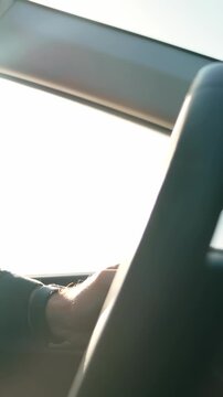 A driver's hand grips a steering wheel in a vehicle, with sunlight filtering through the window. The scene captures the focus of driving and the surrounding environment.