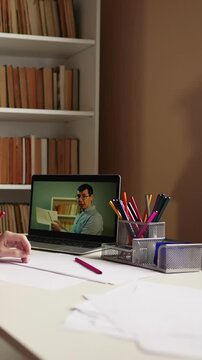 A man reads while looking at a laptop. The desk contains colorful pens and papers, set against a background of bookshelves filled with books.