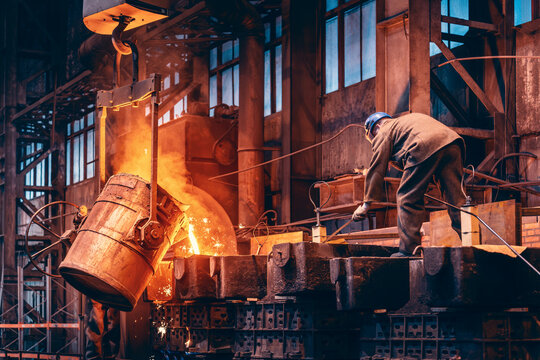 Molten metal pours from massive tilting ladle into casting mold as bright sparks fly during industrial foundry production at metallurgical plant.