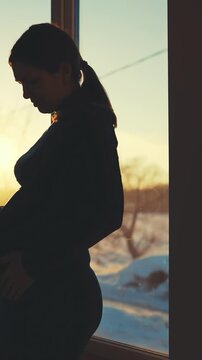 A pregnant woman stands silhouetted against the sunset. She touches her belly while observing the winter landscape outside.