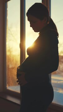 A woman stands by a window, cradling her belly as the sun sets behind her. Soft light creates a silhouette, capturing the intimate moment of motherhood.