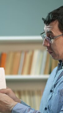 A man in a blue checked shirt holds a book while looking intently. He stands in a room with a shelf of books in the background, showcasing an interest in reading.