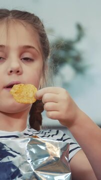 A girl is eating a snack while looking to the side. The girl enjoys a chip from a shiny bag, seated in a well-lit room with greenery in the background.