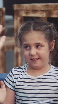 A girl with braided hair smiles while engaging in conversation. She expresses excitement and curiosity with her hands raised in a lively setting.