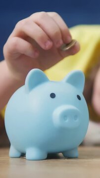 A child's hand reaches towards a blue piggy bank. The child seems focused on the piggy bank as they deposit coins into it.