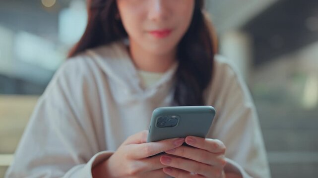 Close up of happy young Asian woman smiling while using smartphone with both hands, typing message and browsing mobile content in casual hoodie, social media, online technology 