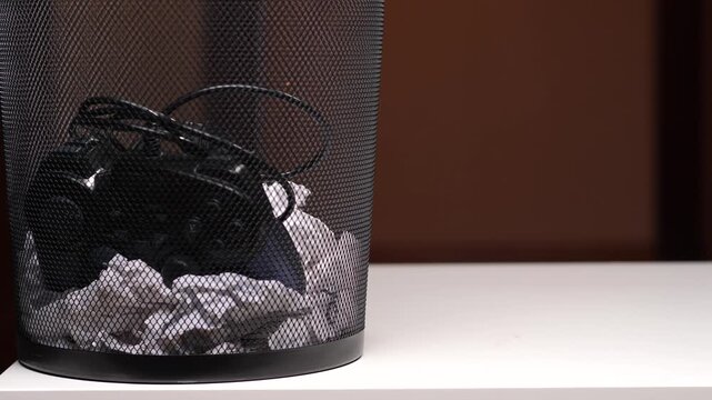 Black wired game controller and crumpled paper tossed in a metal mesh wastebasket, symbolizing gaming addiction, e waste, giving up, digital detox and obsolete technology