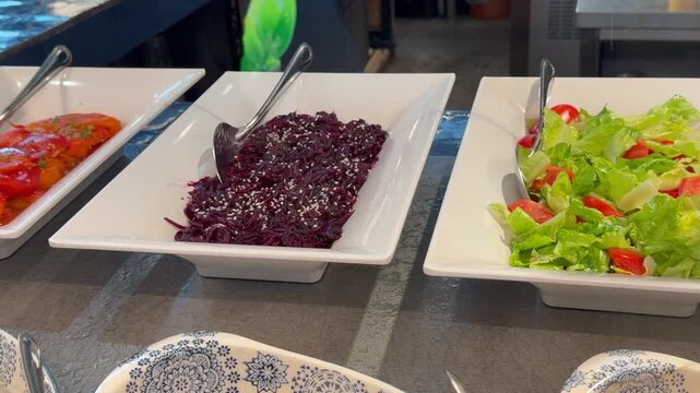 Fresh salads including beetroot with sesame seeds and green lettuce with tomatoes are displayed indoors in white bowls on a buffet counter. Healthy food, catering service, buffet dining.