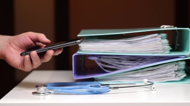 Hand holding smartphone displaying health app, stethoscope and stacked medical folders blurred in background, symbolizing shift from paper patient records to digital ehr efficiency