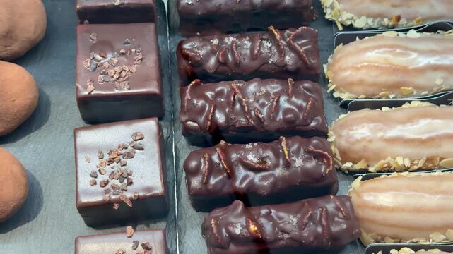 Assorted chocolate pastries and &eacute;clairs with almonds are displayed indoors on a tray at a bakery counter. Sweet desserts, confectionery products, delicious treats.