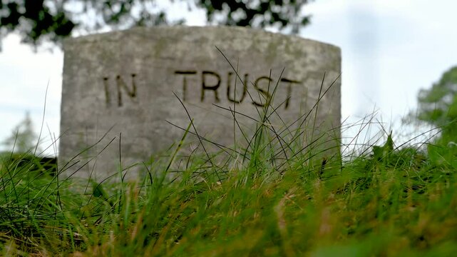 Low angle spooky closeup view of ancient gravestone that reads IN TRUST seen defocused through green grass cemetery under dramatic sky Halloween