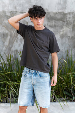Man in a dark t-shirt and denim shorts touching his hair against a concrete wall with tall grass