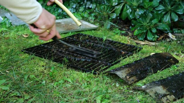 Professional cleaner washing a dirty barbecue grill with a wire brush and a water hose in a garden. The man is scraping off burnt food residue and grease to prepare it for cooking outdoors
