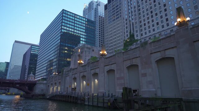 Downtown office towers rise above the Chicago Riverwalk during dusk in Chicago, Illinois. Urban waterfront scene with illuminated riverfront lights, bridge structures, reflections, and modern