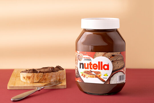 Nutella hazelnut cocoa cream jar beside bread topped with spread on a wooden board, with a spreading knife on a muted red surface against a warm cream color background, with copy space.