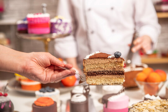 Hand holding a slice of layered chocolate sponge cake on a server, with decorated desserts in a pastry workshop.