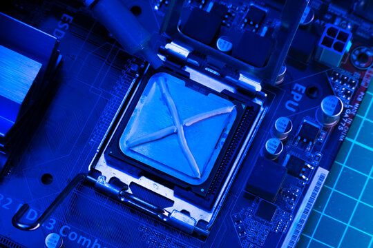 neon blue illuminated pc motherboard with a syringe adding thermal compound in an x shape onto the cpu processor