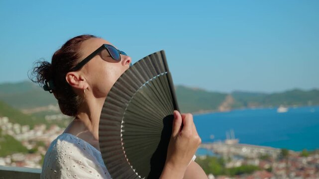 Woman suffering from summer heat using fan. Exhausted woman in sunglasses using a hand fan to cool down on a hot summer day, sitting on a balcony with a beautiful sea view. Extreme heatwave concept