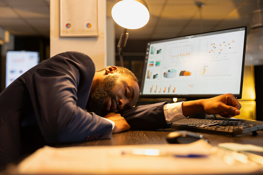 Tired Black startup employee falls asleep at his desk after hours, struggling with a heavy workload and burnout from the pressure of late-night work.