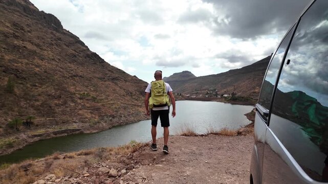 Man with backpack pauses by his parked van at a mountain-lake viewpoint, soaking in tranquil scenery during a solo road-trip hike for wellness, freedom and adventure