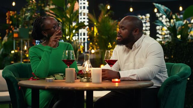 Cheerful couple has fun chatting during an entertaining evening in the city center while smiling partners enjoy wine at a romantic rooftop restaurant.
