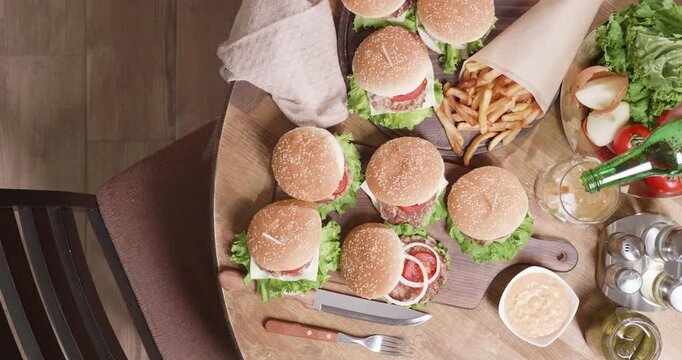 Beer, pouring and burgers in a top-down pub dining scene as a green bottle pours beer into a glass beside sesame-bun cheeseburgers, French fries, fresh tomatoes and condiments for casual meals.