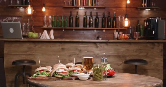 Pub, burgers and hospitality in a wide shot of a warmly lit wooden bar with a prepared cheeseburger platter, fries, beer and condiments on the foreground table for restaurant ambience and menu use.