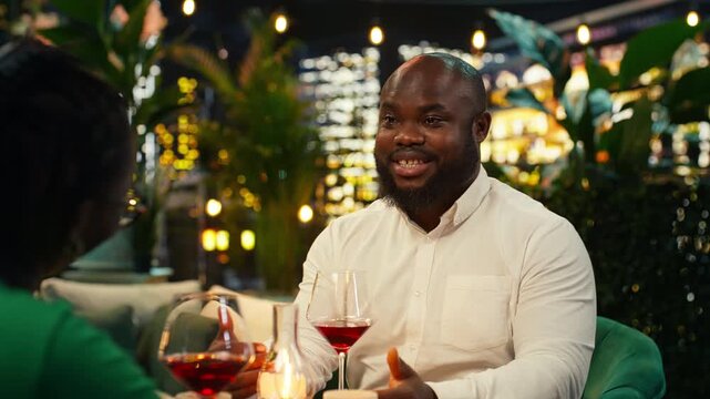 Smiling boyfriend chats with his girlfriend in an outdoor rooftop lounge, while the cheerful man shares a story with his partner during a candlelit restaurant date among lush plants at night.