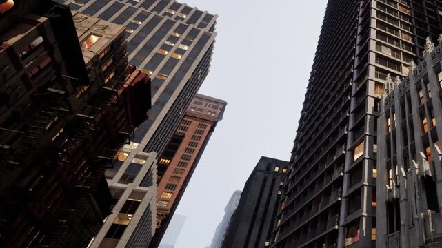 Tall commercial buildings rise into the bright sky above a city street during the day as sunlight hits the glass windows of the towering structures that define the busy urban skyline of New York City