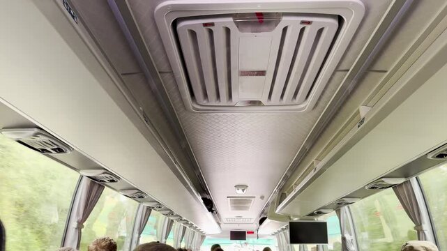 A roof vent blows cool air inside a moving bus ceiling. Passengers enjoy a smooth journey past green trees. The clean interior creates a calm and comfortable feeling.