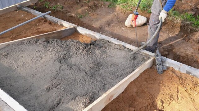 Construction worker compacts wet concrete in foundation formwork at outdoor residential site, with soil trench, timber edging, and bright daylight showing ongoing home development.