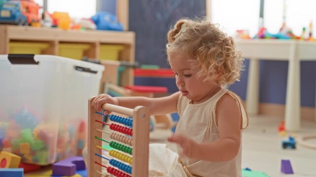 Little blonde girl seated on foam mat pushes multicolored beads on a wooden abacus with her hand in a bright daycare playroom; curiosity.