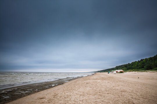 Jurmala beach in Dubulti, Latvia, beneath an overcast sky. Baltic Sea waves wash onto the sandy shore, with coastal woodland and a beach building extending along the quiet seaside resort waterfront.