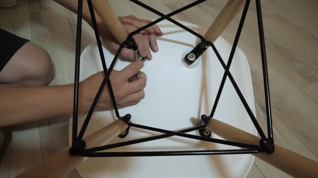 Close-up overhead view shows male hands tightening bolts and aligning connectors beneath white round seat during chair assembly, with careful rotation, fitting and support placement