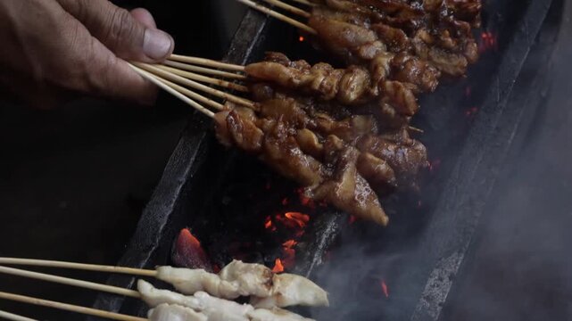 Cinematic close-up video of a hand slowly turning chicken satay skewers over glowing charcoal. Smoke drifts upward as the meat sizzles, lit by warm, dramatic light.