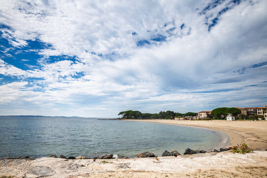 Panoramic view of Plage de Miramar in La Londe les Maures, France, on the French Riviera. A curved sandy beach borders the Mediterranean Sea, with coastal trees and buildings beneath a cloud-filled sk