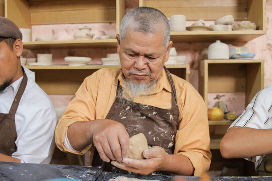 Focused Elderly Craftsman Wearing An Apron Molding Raw Material At His Table