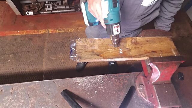 Electric stapler fastens wood plank during workshop use. Worker presses power tool onto board clamped in metal vise, creating practical fastening setup with focused precision for secure assembly.