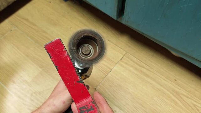 Hands test impact driver with wire brush in workshop demo. Red abrasive strip moves near spinning attachment as tool runs, with both hands guiding setup over wood floor during surface work.