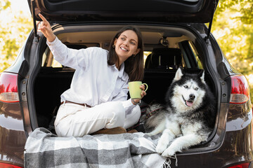 Road trip adventure and travel lifestyle, happy woman with husky dog sitting in car trunk pointing at view with coffee cup during summer vacation © Pixel-Shot
